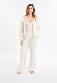 Ensemble de vêtements d'intérieur beige comprenant un sweat à capuche zippé avec des poignets élastiques, un body à col en V, et un pantalon large à coupe décontractée. Texture douce.