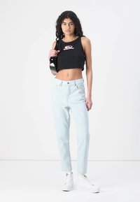 Top corto nero con logo stampato, jeans a vita alta blu chiaro e sneakers bianche con piattaforma. Il modello si trova su uno sfondo neutro.