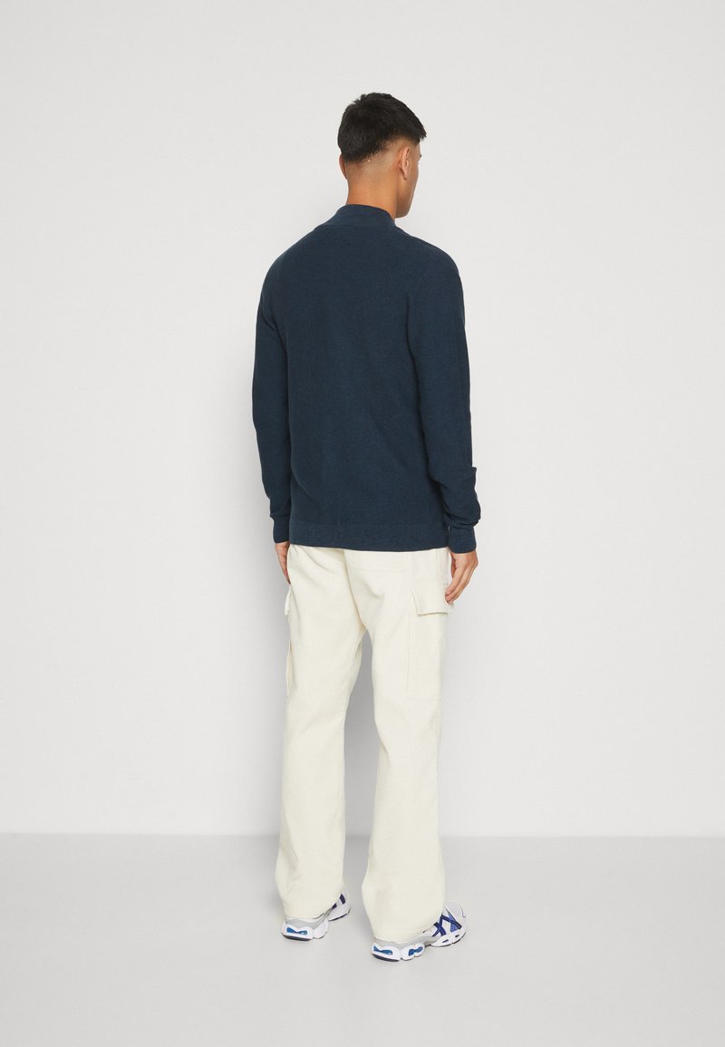 Granatowy sweter z dzianiny o prążkowanej fakturze, w zestawieniu z białymi spodniami cargo. Strój posiada długie rękawy oraz luźny krój.