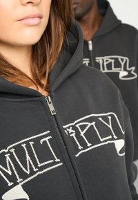 Sudadera negra con cremallera y la palabra "MULTIPLY" en texto blanco bordado, fabricada en un material suave, con una capucha grande y una cremallera plateada con un tirador triangular.