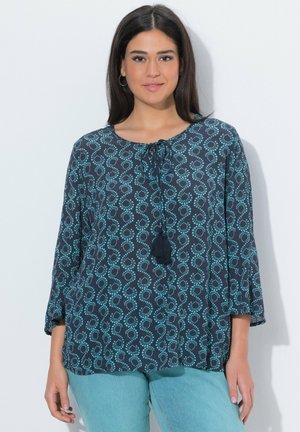 Femme portant un chemisier bleu marine avec des motifs en boucles pointillées turquoise et des liens à pompons, associée à un jean bleu clair, debout contre un mur blanc.