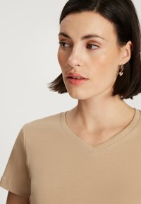Beżowa koszulka z dekoltem w serek uszyta z miękkiego materiału, w taliowanym kroju, z krótkimi rękawami. Posiada gładką fakturę i subtelne detale szycia.