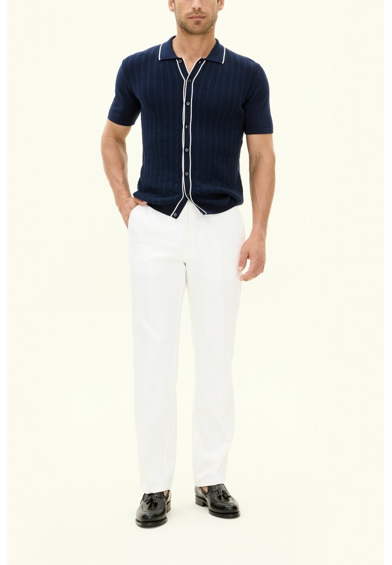 Marineblauwe ribgebreide T-shirt met korte mouwen en witte afwerking, gecombineerd met witte broeken en zwarte schoenen. Zichtbare knopen aan de voorkant.