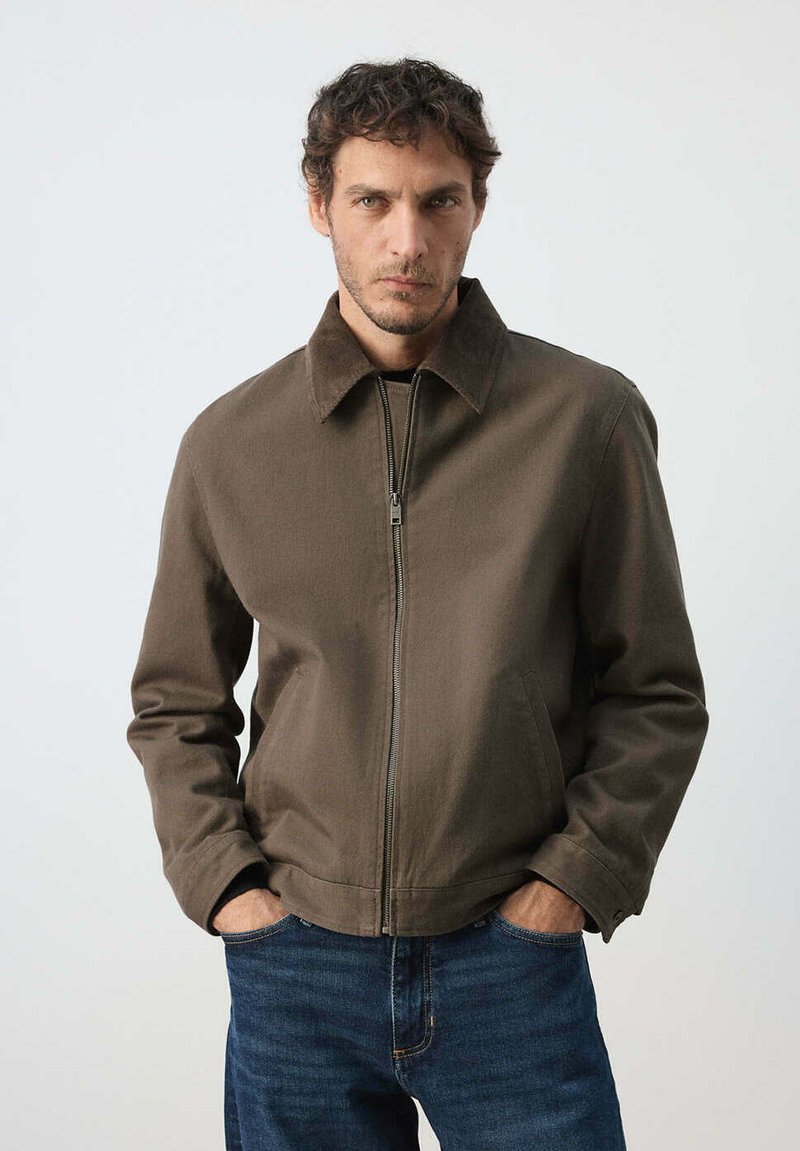 Homme aux cheveux courts et bouclés, portant une veste marron zippée et un jean bleu, debout les mains dans les poches devant un fond uni.