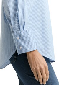 Light blue striped button-up shirt with small metallic accents. Features a cuff with two white buttons and a slightly longer back hem.