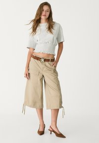 Svetlý šedý crop top so stiahnutým pásom, skombinovaný s béžovými voľnými cargo nohavicami s vreckami a sťahovacími šnúrkami; hnedý opasok so sponou v tvare hviezdy.