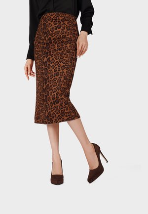 Donna che indossa una gonna a tubino marrone con stampa leopardata, top nero e scarpe con tacchi alti marrone scuro, in piedi con una gamba leggermente sollevata.