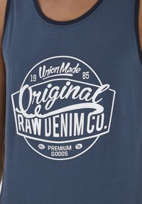 Sötétkék pamut ujjatlan felső, amelyen kör alakú fehér grafika látható. A grafika tartalmazza a következő szövegeket: "Union Made", "Original" és "Raw Denim Co." különböző betűstílusokban.