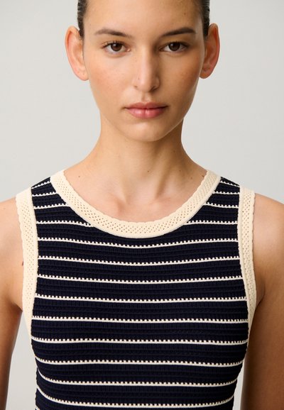 Frau trägt ärmelloses, schwarz-creme gestreiftes Stricktop mit cremefarbenem Besatz, dargestellt vom mittleren Brustbereich bis zum Kopf vor einfarbigem Hintergrund.