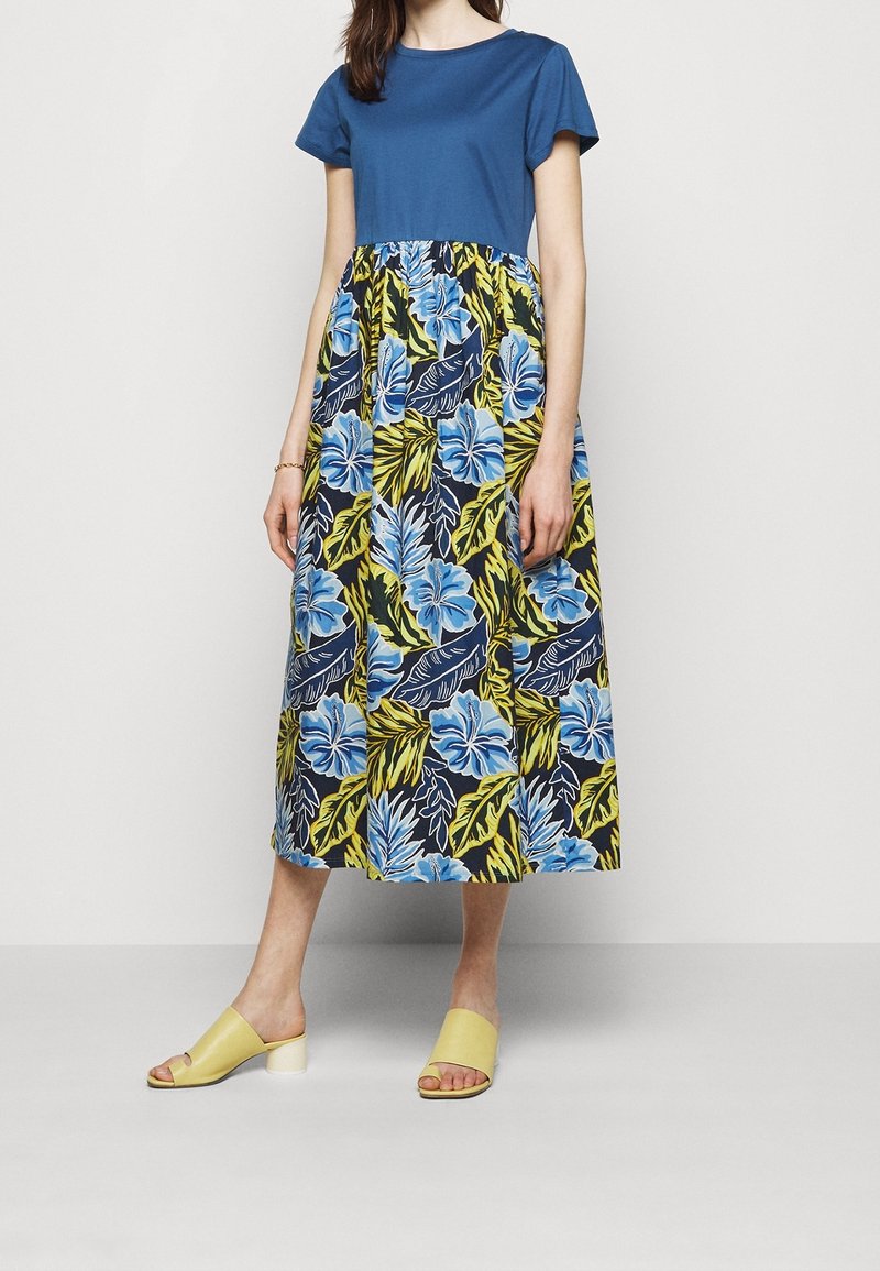 Femme portant une robe à manches courtes bleue avec une jupe à motif floral jaune, bleu et noir, et des sandales jaunes à talon bas.