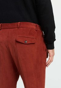 Rote Cordhosen mit vertikaler Rippstruktur, ausgestattet mit einer Gesäßtasche mit Klappe und Knopfdetail, kombiniert mit einem schwarzen Langarmshirt.