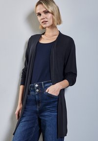 Svart stickad cardigan med öppen framsida och långa ärmar, bärs över en marinblå topp. Matchas med mörkblå jeans med hög midja och fickor.