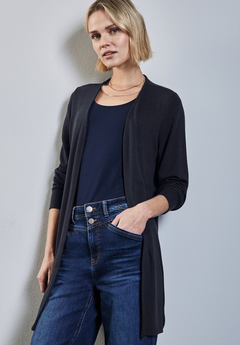 Svart stickad cardigan med öppen framsida och långa ärmar, bärs över en marinblå topp. Matchas med mörkblå jeans med hög midja och fickor.