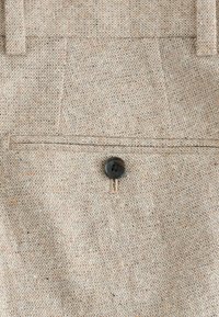 Light brown textured fabric with a subtle speckle pattern. Features a back pocket with a single black button closure.