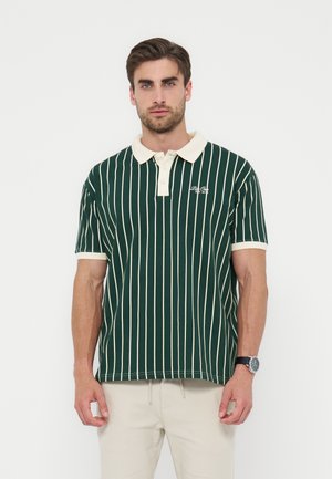 Mann, der ein dunkelgrünes kurzärmeliges Polo mit weißen vertikalen Streifen, cremefarbenem Kragen und hellen Hosen trägt und vor einem weißen Hintergrund steht.