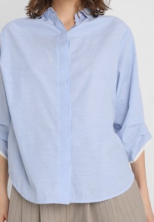 Blouse bleu clair à manches longues avec patte de boutonnage cachée, col, et manches retroussées portée avec un pantalon plissé beige.