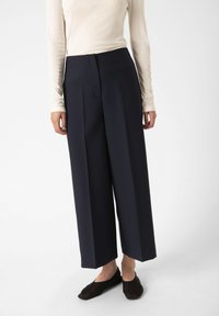 Pantaloni a gamba larga blu navy con pieghe frontali, abbinati a un top a maniche lunghe beige chiaro. Texture liscia, design minimalista, senza motivi visibili.