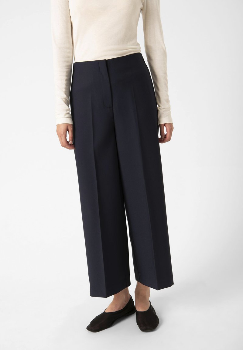 Pantaloni a gamba larga blu navy con pieghe frontali, abbinati a un top a maniche lunghe beige chiaro. Texture liscia, design minimalista, senza motivi visibili.