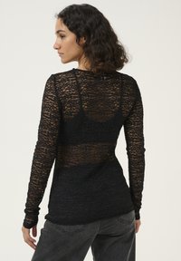 Black long-sleeve top made of sheer, textured fabric with a layered design. Features fitted sleeves and an open back. Worn with black jeans.