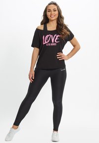 Black off-shoulder T-shirt with pink text "LOVE IS THE ANSWER," paired with black leggings. Model stands in white sneakers on a neutral background.