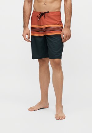 Homme portant un short de bain mi-long avec des rayures horizontales rouges, orange et noires, debout pieds nus sur un fond blanc.