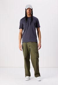 Vyrų apranga, sudaranti prigludusią tamsiai mėlyną marškinėlį ir laisvus alyvų spalvos krovinius, turinčius kelias kišenes. Papildyta šviesiai pilku kepurėle ir baltais sportbačiais.