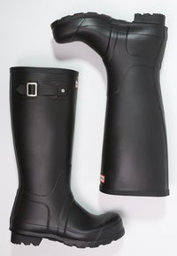 Botas de lluvia de goma negra con suelas texturizadas, un acento de hebilla en la parte superior y un acabado suave; una bota está en posición vertical y la otra está en ángulo.