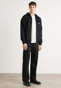 Svart zip-up hettegenser med kengurulommer, parret med løse svarte cargo-bukser med sidelommer og sølv-farge snaps knapper. Svart sko.