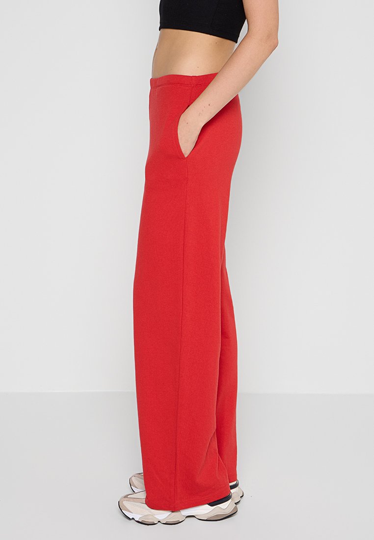 Pantalons de survêtement rouge à jambes larges avec une texture douce, dotés d'une taille élastique et de poches latérales, associés à des baskets neutres.