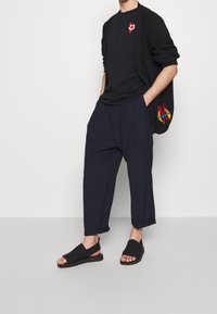 Černý cardigan s červenými a žlutými vyšitými akcenty, černé tričko, volné námořnické kalhoty a černé sandály na klouzání s texturovanými podrážkami.