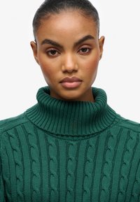 Zielony sweter o grubym splocie z wysokim, prążkowanym golfem i teksturowanym wzorem w warkocze. Gładki materiał z dopasowanym krojem.