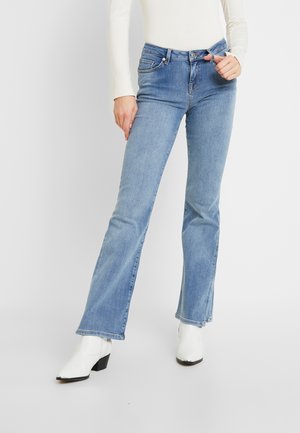 Vrouw draagt lichtblauwe bootcutjeans, wit shirt met lange mouwen en witte enkellaarzen, staand tegen een eenvoudige lichte achtergrond.