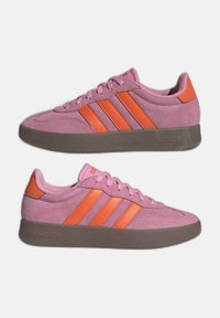 Ett par rosa mocka Adidas-sneakers med orange ränder och detaljer, bruna gummisulor och snören, visade i sidoprofil mot vit bakgrund.
