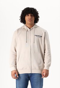 Beige zip-up hoodie met trekkoordkap, vo pockets en tekstlogo op de linkerborst. Gecombineerd met blauwe denim jeans.