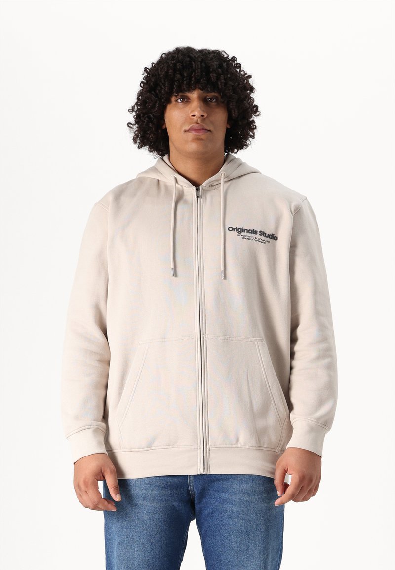 Beige zip-up hoodie met trekkoordkap, vo pockets en tekstlogo op de linkerborst. Gecombineerd met blauwe denim jeans.