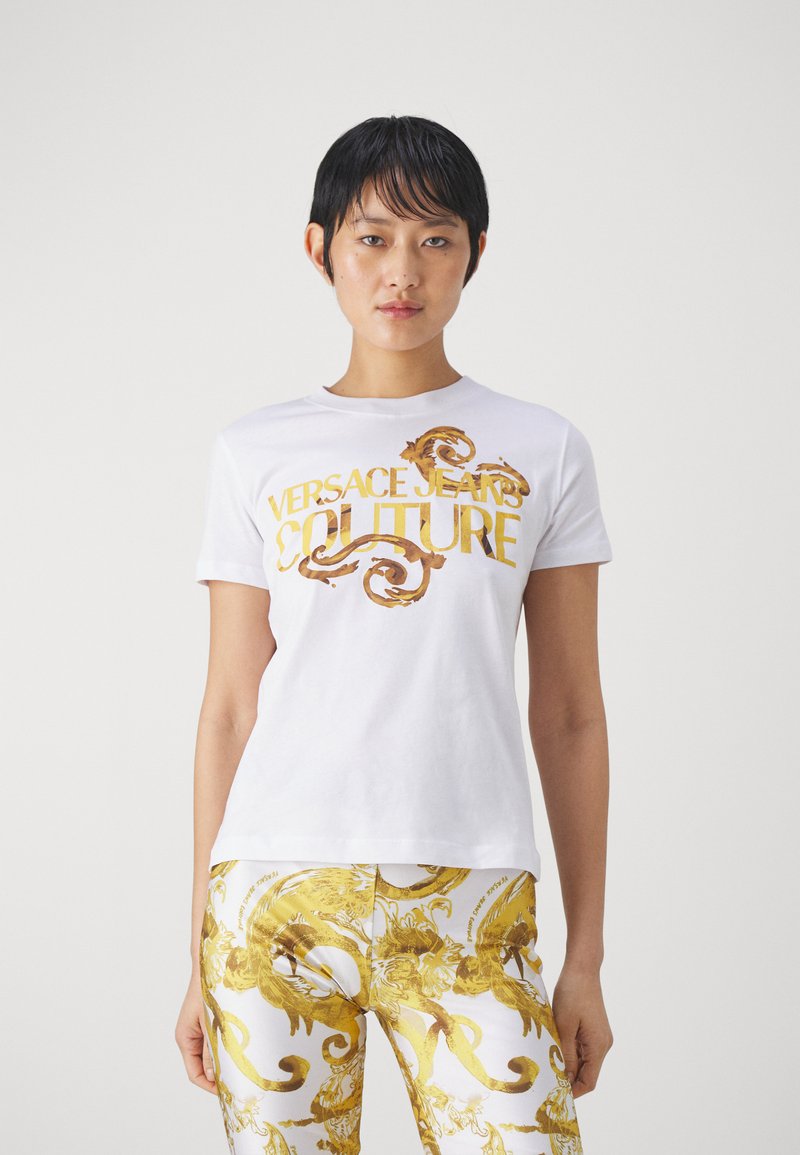 Λευκό βαμβακερό T-shirt με χρυσό κείμενο "VERSACE JEANS COUTURE" και στρογγυλεμένα σχέδια. Το μοντέλο το φορεί με παντελόνι με χρυσά σχέδια.