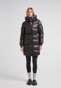 Mujer vistiendo un abrigo acolchado negro con cinturón y collar alto, leggings negros, calcetines grises y zapatos negros de tipo slip-on, de pie contra un fondo sencillo.