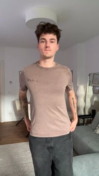 Beżowa koszulka z krótkim rękawem o wyblakłym wyglądzie z logo na lewej piersi, zestawiona z ciemnymi spodniami. Gładka tkanina, swobodny krój.