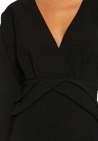 Robe noire enveloppante avec un décolleté en V profond, des manches longues, un tissu texturé et un design avant torsadé pour une touche de détail supplémentaire.