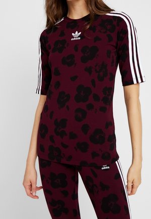 Kobieta ubrana w bordowy top i legginsy Adidas z czarnym kwiatowym wzorem i białymi trzema paskami na rękawach i bokach spodni.