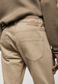 Beige Denim-Hosen mit mittelhohem Bund, zwei Gesäßtaschen und einem Lederetikett. Die Textur wirkt glatt; subtile Nähte sind sichtbar.