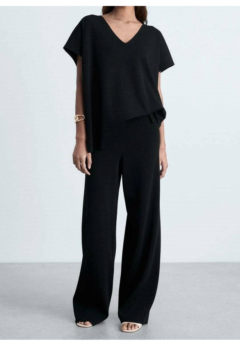Femme portant un haut noir ample à col en V et un pantalon noir à jambes larges, des sandales beige à bout ouvert et un bracelet en or au poignet gauche.