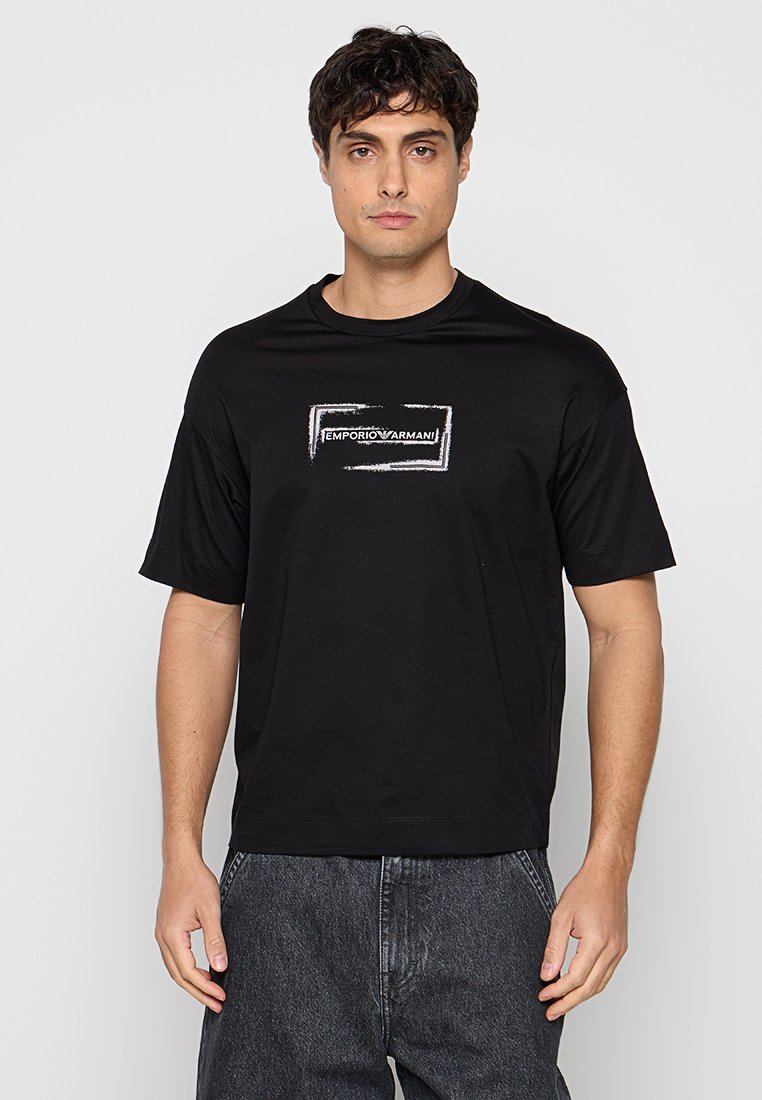 Hombre con camiseta negra de Emporio Armani y vaqueros grises, de pie frente a un fondo liso de color gris claro.