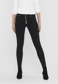 Jeans skinny negros de cintura alta con un cierre de cinco botones, bolsillos delanteros y una textura de mezclilla suave. Combinados con botas de tobillo negras.