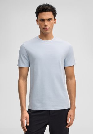 Mann mit kurzen lockigen Haaren, der ein hellblaues, strukturiertes Kurzarmhemd und dunkle Hosen trägt, vor einem einfarbigen grauen Hintergrund steht.