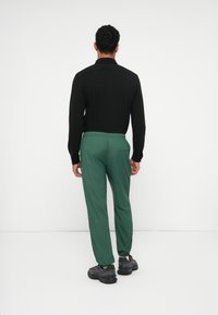 Černá košile s dlouhými rukávy a rovného střihu, spojená se zelenými kalhotami na gumu. Outfit doplňují černé sportovní boty.