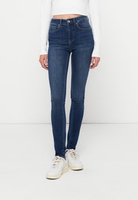 Sötétkék farmerből készült, magasított derekú, skinny fazonú farmernadrág, elülső zsebekkel, cipzáras nyitással és sima textúrával. Fehér sportcipővel viselve.