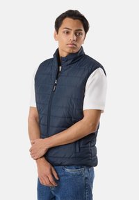 Ung mand med mørkt hår iført en marineblå quiltet lynlåsvest over en hvid kortærmet skjorte og blå jeans, stående med armene krydsede.