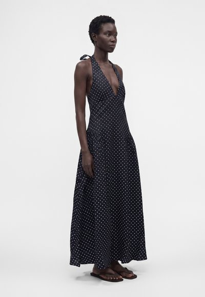 Γυναίκα με μαύρο μακρύ φόρεμα με halter neck και λευκά πουά, στέκεται με καφέ σαγιονάρες σε απλό φόντο.