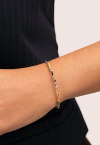 Armband med pärlor i grå och svarta stenar, blandat med detaljer i guldfärgad metall, bärs på ett handled mot ett mörkt, randigt tyg.